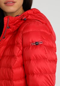 Vrouw in een felrode gewatteerde pufferjack met capuchon, voorzien van een klein rechthoekig logo-opdruk op de bovenste mouw.