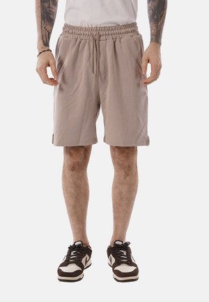 Uomo con pantaloncini beige con vita elastica e coulisse, t-shirt bianca, scarpe da ginnastica nere e bianche, in piedi con le mani rilassate lungo i fianchi.