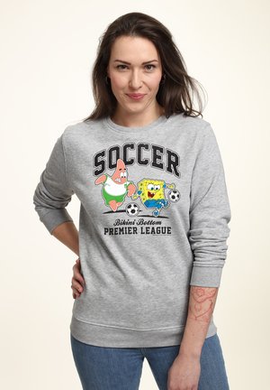 Frau trägt grauen Pullover mit SpongeBob und Patrick, die Fußball spielen, mit dem Text „Bikini Bottom Premier League.“