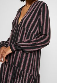 Černé šaty s červenými a bílými vertikálními pruhy, s hlubokým výstřihem do V, dlouhými rukávy a vrstveným volánkovým lemem. Texturovaná tkanina s elastickými manžetami.