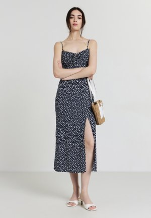 Robe midi florale navy avec des bretelles fines ajustables, une fente sur le côté et un corsage ajusté. Associée à des sandales blanches à talons carrés et un sac en osier.