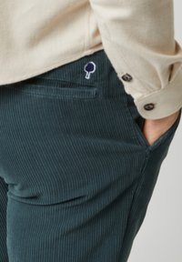 Donkergroene corduroybroek met een gestructureerd oppervlak en smalle ribbels, voorzien van een klein geborduurd boomlogo nabij het achterzakje.