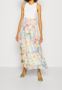 Maxime dress with a fitted white bodice and a layered, colorful patterned skirt featuring abstract drawings; paired with green strappy sandals.