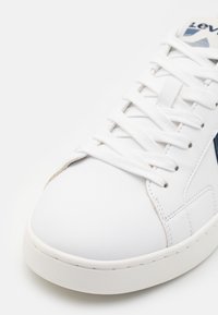 Białe sneakersy z gładką, syntetyczną cholewką, z granatowym logo i białymi sznurowadłami. Podeszwa jest okrągła i lekko podniesiona.
