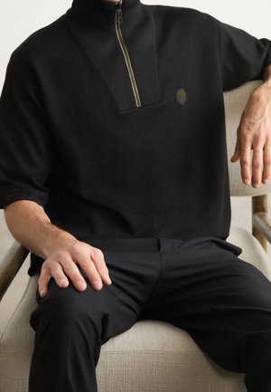 Zwarte half-zip pullover met een gestructureerde kraag, gemaakt van zachte stof. Kenmerkt zich door een klein logo-detail en een relaxte pasvorm. Gepaard met zwarte broeken.