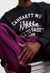 Persona che indossa un felpa nera con figure evolutive bianche e la scritta "Carhartt WIP Back to Basics Est. 1989", mentre tiene in mano un paio di pantaloni piegati viola.