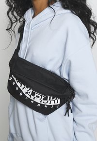 Černá ledvinka se zipovou kapsou, s bílým logem. Vyrobena z odolné látky, nošena přes hruď na světle modré mikině.