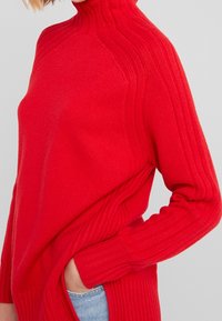Pull à col roulé en tricot rouge avec des détails côtelés, des manches longues et une coupe décontractée. Fabriqué en matériau doux et texturé.
