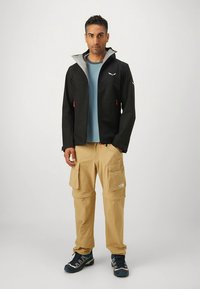 Black waterproof jacket with gray accents, worn over a light blue shirt and beige cargo pants, complemented by blue hiking shoes.