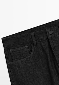 Sorte denimbukser med et klassisk design, inklusive knaplukning, bæltestropper og en forlomme, der viser en tekstureret stofoverflade.