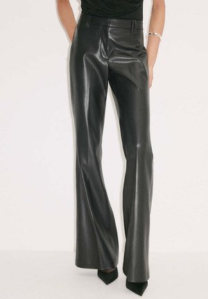 Femme portant un pantalon large en cuir synthétique noir taille haute, des chaussures noires à bout pointu et un haut noir.