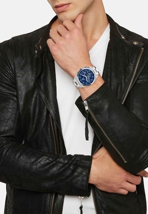 Orologio argento con quadrante blu, dotato di più quadranti, indossato al polso. La mano della persona poggia su una giacca di pelle nera.