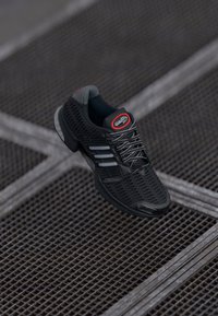 Chaussure de sport noire avec une tige en maille, des côtés texturés et des accents réfléchissants ; dispose de lacets gris et d'un logo rouge proéminent.