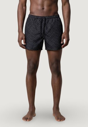 Hombre atlético con pantalones cortos negros con cordón y un patrón diagonal repetitivo de texto "CHANGE YEAR", de pie descalzo frente a un fondo liso.