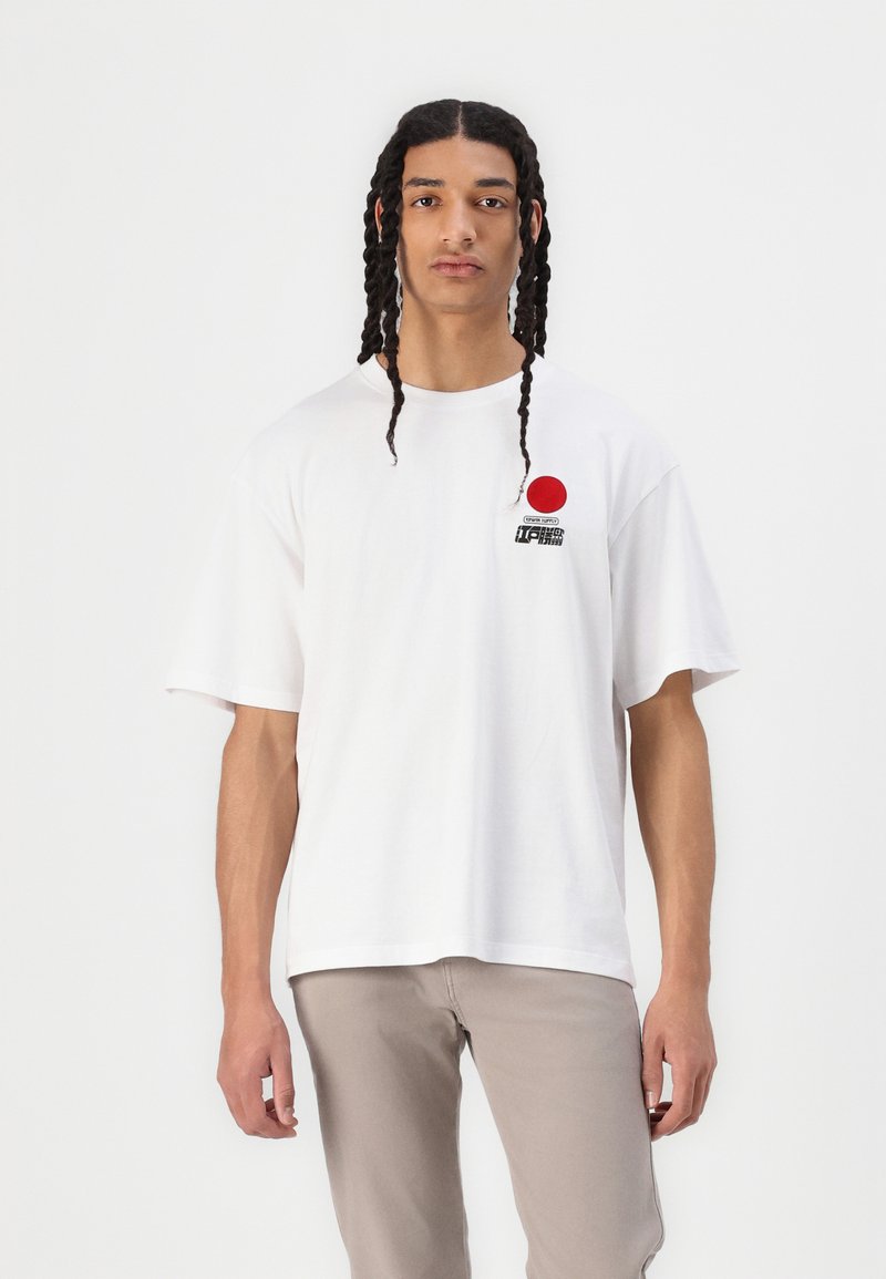 White short-sleeve t-shirt featuring a red circle and graphic print on the left chest, made of smooth cotton fabric. Model wearing beige pants.