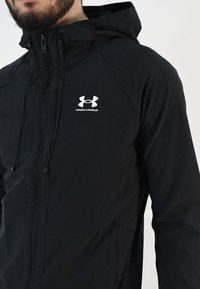 Chaqueta negra con capucha hecha de una tela ligera. Cuenta con un cierre de cremallera y un logo de Under Armour en la parte izquierda del pecho.