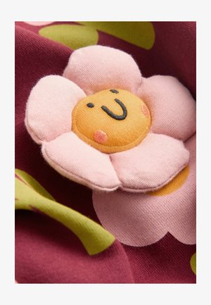 Pluchen bloemenapplicatie met roze bloemblaadjes en een gele kern, met een glimlachend gezicht, tegen een gestructureerde bordeauxrode en groene achtergrond.