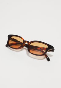 Ochelari de soare tortoise maro cu lentile rectangle portocalii, cadru gros și accente negre pe tempă. Design în culori neutre.