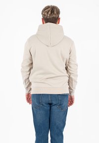 Felpa beige chiaro con un ampio cappuccio, polsini e orlo a coste. Indossata con jeans in denim blu, vista da dietro. Texture liscia, design casual.