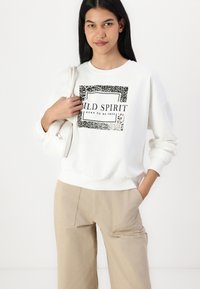 Witte sweatshirt met "WILD SPIRIT" grafiek, rand met dierenprint. Heeft ribgebreide manchetten en tailleband. Draagt met beige broeken. Kleine witte tas.
