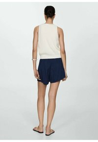 Cremefarbener, gestrickter ärmelloser Top, kombiniert mit dunkelblauen Shorts, die einen elastischen Bund und eine legere Passform haben. Sandalen runden den Look ab.
