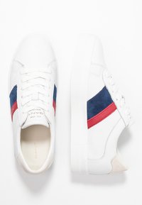 Białe skórzane sneakersy z granatowymi i czerwonymi zamszowymi paskami, płaskimi sznurowadłami oraz teksturowaną gumową podeszwą z beżowym akcentem na pięcie.