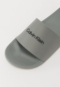 Sandale de plajă din cauciuc gri, cu o curea lată care prezintă logo-ul "Calvin Klein" în negru. Tălpic texturat și suprafață a tălpii netedă.