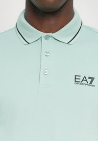 Polo verde claro com um acabamento texturizado, três botões e colarinho com riscas pretas. Apresenta o logótipo EA7 Emporio Armani no lado esquerdo do peito.