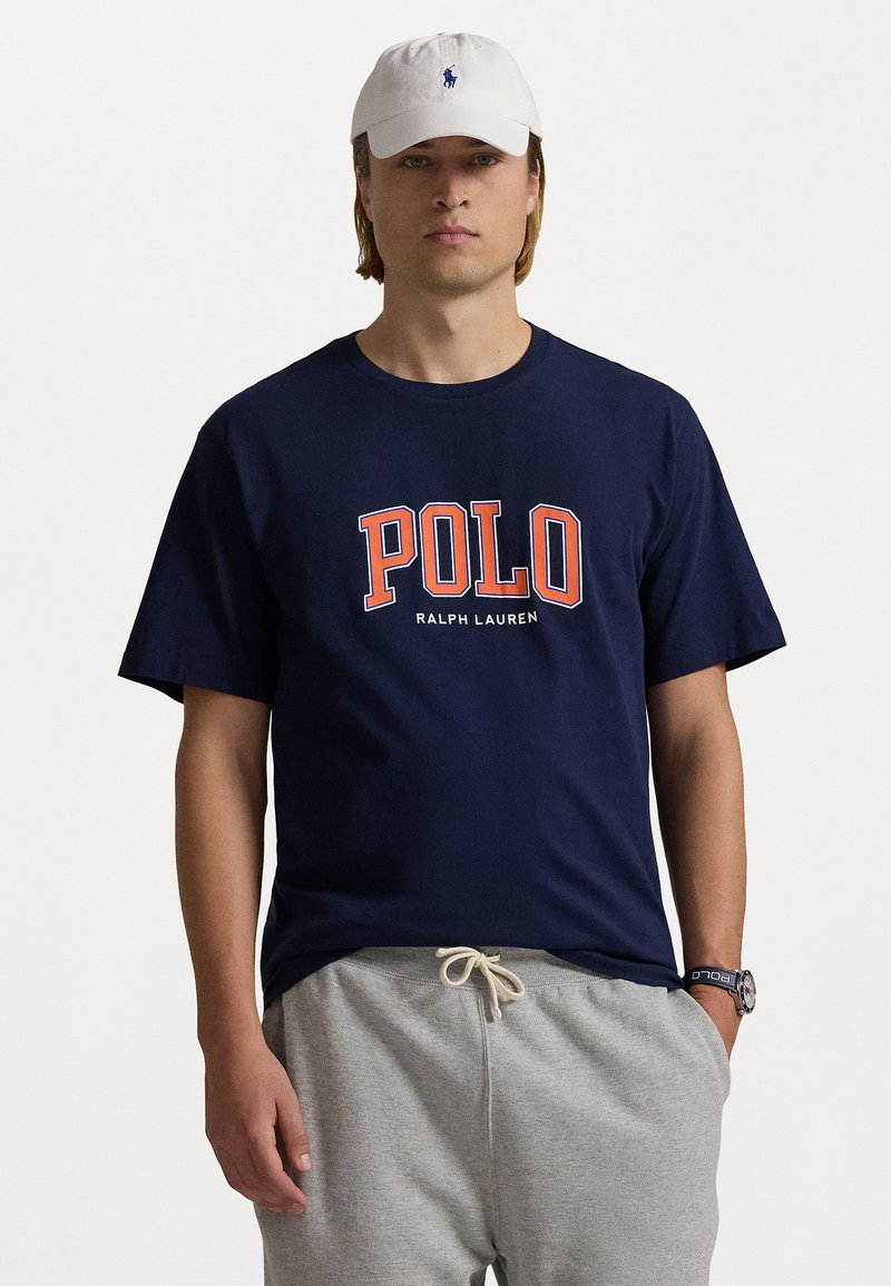 Jūras spēku t-kreklis ar "POLO" un "RALPH LAUREN" uzrakstiem oranžā un baltā krāsā. Apvienots ar pelēkiem sporta biksēm un baltu cepuri ar logotipu.
