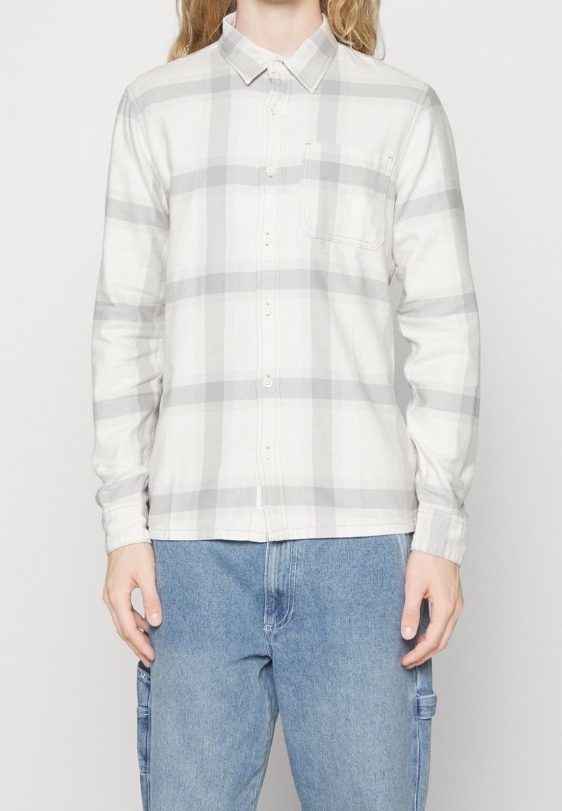 Camicia bianca con bottoni, modello a scacchi grigi, maniche lunghe e taschino sul petto, realizzata in un tessuto morbido e testurizzato.