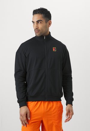 Jachetă neagră cu fermoar, guler înalt, având un logo mic multicolor pe partea stângă a pieptului; purtată cu pantaloni scurți strălucitori de culoare portocalie.