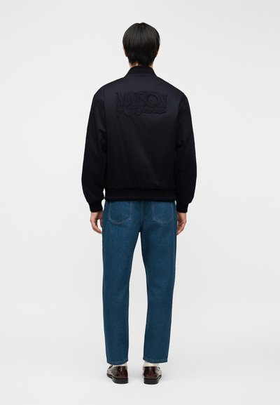 Person steht mit dem Rücken zur Kamera, trägt eine schwarze Jacke mit der Aufschrift "Maison Kitsuné" auf dem Rücken, blaue Jeans und dunkle Schuhe.