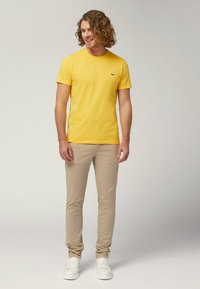 T-shirt di cotone gialla con collo rotondo e maniche corte. Pantaloni slim-fit di color beige chiaro. Modello che indossa sneakers bianche, capelli leggermente ricci.