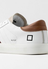 Hvide lædersneakers med en brun læderhælkappe, der har en tekstureret indvendig, sort logo, og en hvid gummisål.
