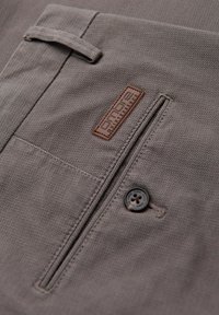 Gros plan d'un pantalon en tissu gris montrant une poche arrière avec un bouton, une passants de ceinture et une étiquette de marque marron « ombre ».