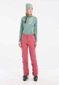 Giacca zip verde chiaro con una finitura testurizzata, abbinata a pantaloni impermeabili rosa; stivali da sci neri completano l'outfit.