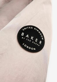 Svart cirkulär etikett på ljusrosa tyg, med texten "BAKER by TED BAKER" och "UNITED KINGDOM LONDON" i vitt. Slät textur.