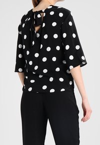 Blouse noire à pois blancs, manches courtes, dos en trou de serrure avec détail nœud, tissu lisse, coupe décontractée. Associée à un pantalon noir.