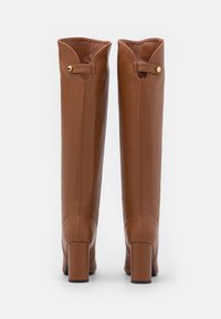 Par de botas altas de cuero marrón con tacones cuadrados, puños doblados y acentos en botón dorado, mostradas desde atrás sobre un fondo blanco.