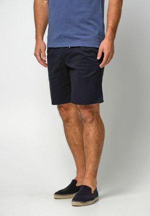 Marineblaue Freizeit-Shorts mit flacher Vorderseite, Seitentaschen und entspannter Passform, kombiniert mit dunklen Slips und einem blauen T-Shirt.