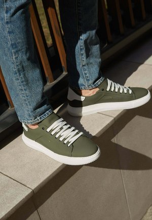 Olijfgroene sneakers met witte veters, rubberen zolen en een contrasterend hakontwerp. Soepele textuur met stikseldetails.