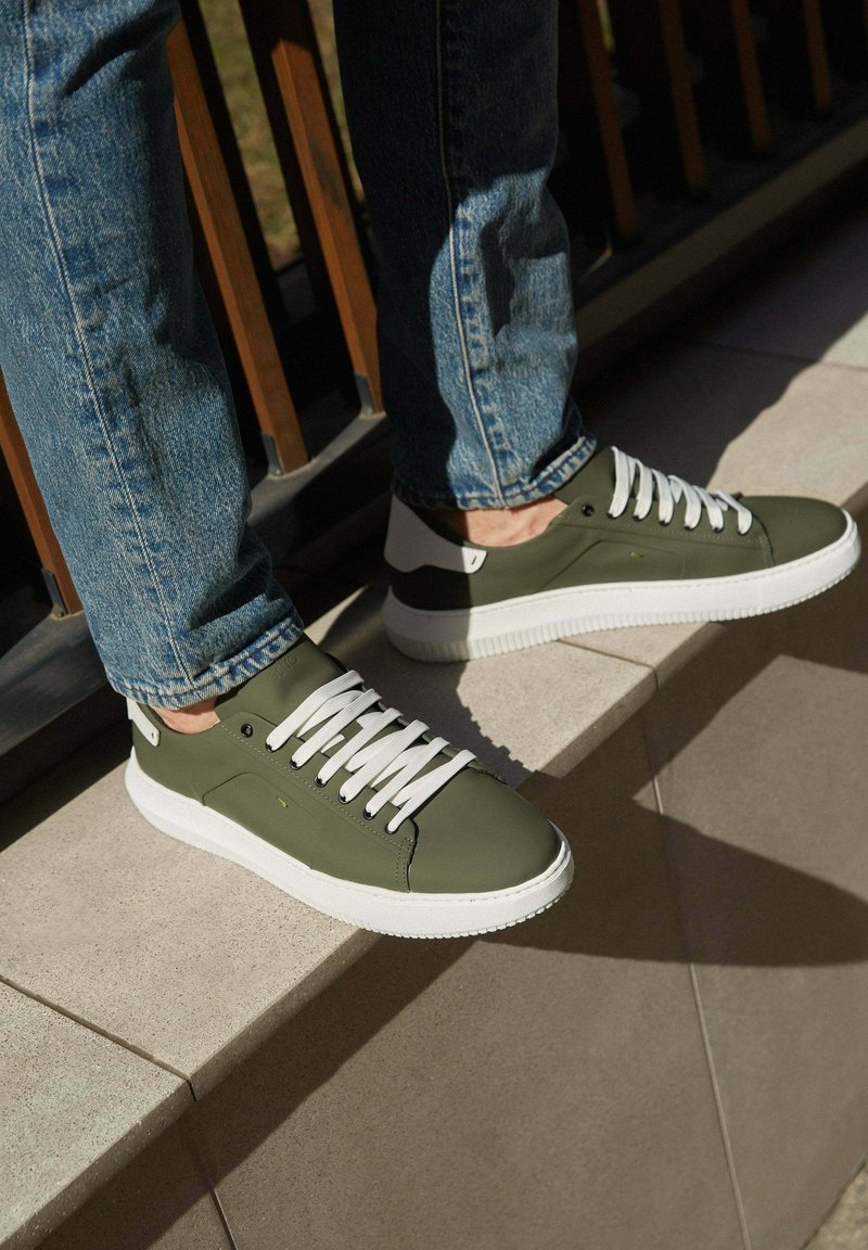 Olivově zelené tenisky s bílými tkaničkami, gumovými podrážkami a kontrastním designem paty. Hladký povrch s švy.