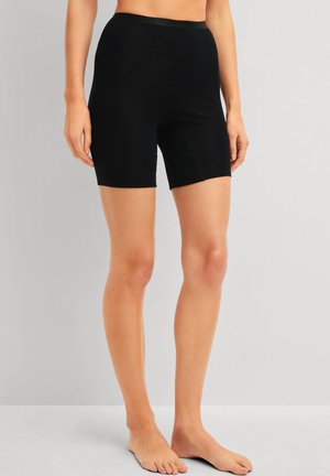 Femme portant un short noir ajusté taille haute, debout pieds nus devant un fond gris clair uni.