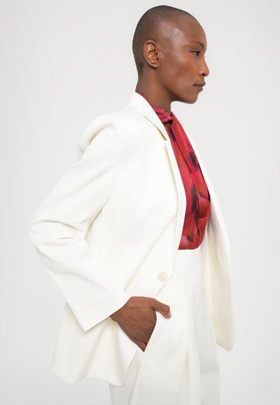 Witte blazer met schoudervulling, sluiting met één knop en grote zakken, gedragen over een blouse met een rood en zwart patroon. Gladde textuur.