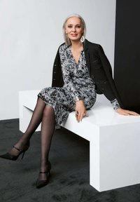 Schwarz-weiß gemustertes Kleid mit langen Ärmeln, kombiniert mit einem schwarzen strukturierten Blazer. Das Model sitzt auf einer weißen geometrischen Bank. Schwarze High Heels.