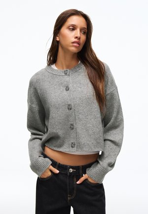 Jeune femme portant un cardigan court gris boutonné et un jean noir, les mains dans les poches, regardant sur le côté sur un fond blanc.
