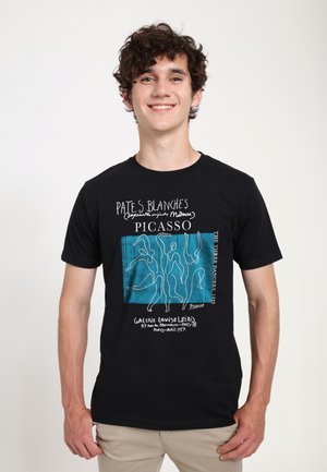 Ung mand smilende iført sort T-shirt med Picassos "De Tre Dansere" 1925 kunstværk design, stående mod en ensfarvet hvid baggrund.
