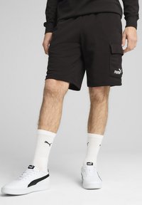 Black cotton shorts with side cargo pocket and white Puma logo. Paired with white sneakers and ribbed white socks featuring black logo.