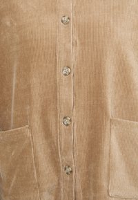 Beige corduroy-takki, jossa on pystysuorat urat, viisi nappia keskellä ja kaksi etutaskua alhaalla.