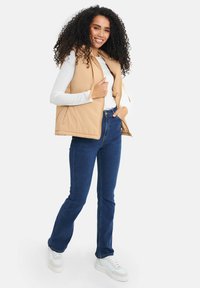 Beige puffer-väst med hög krage, dragkedja och snören, bärs över en vit långärmad tröja, ihop med mörkblå utsvängda jeans och vita sneakers.